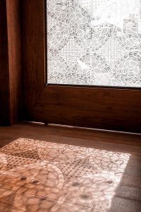 Porta de vidro com renda renascença com sol de fora pra dentro, gerando sombras no piso de madeira.